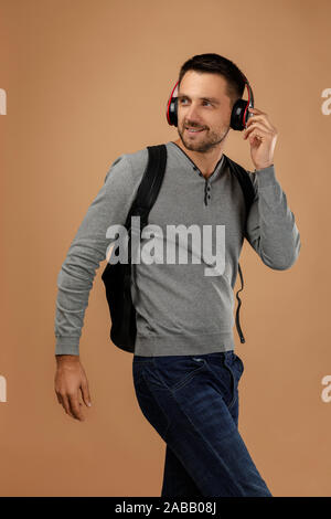 handsome young man in headphones with backpack listens to music isolated on beige background. Stock Photo