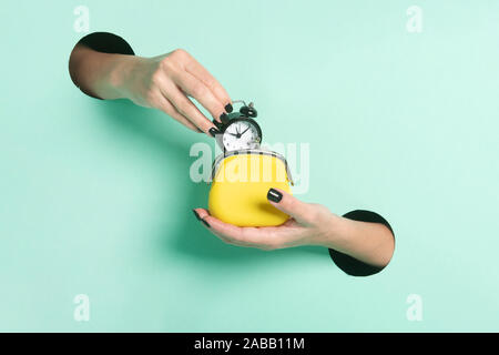 Female hands hold alarm clock and wallet through a hole on neon mint background. Time is money concept. Stock Photo