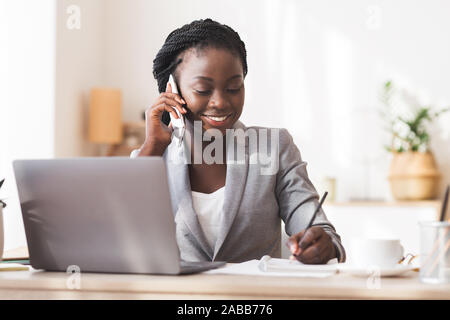 Afro Secretary Taking Notes And Talking On Cellphone In Office Stock Photo