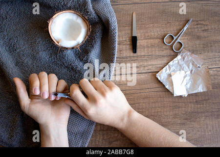 Hand care, folk hand care. Manicure with coconut oil, manicure tools: scissors, nail file. Foil for removing gel polish on wooden background. Beauty a Stock Photo