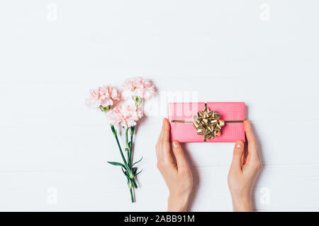 Female hands holding pink gift box with golden bow next to fresh flowers bouquet on white background, flat lay. Stock Photo