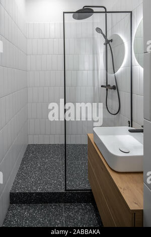 Stylish luminous bathroom with tiled white walls and textured dark gray floor. There is a black shower, glass partition, round mirror with backlight, Stock Photo
