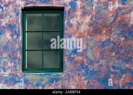 Window with green wood trim set in a weathered plaster wall with a distressed paint style in vibrant colors of blue, purple, and pink Stock Photo
