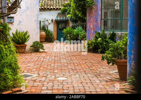 Picturesque garden courtyard with potted plants and enclosed by rustic plaster walls in vibrant hues of purple, pink, and blue and cobbled with brick Stock Photo