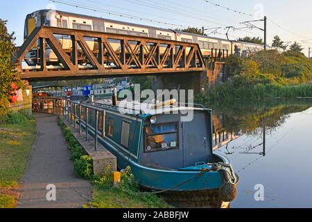 Stansted Express passenger train approaching Ely railway station on steel bridge above River Great Ouse narrowboat & towpath East Anglia England UK