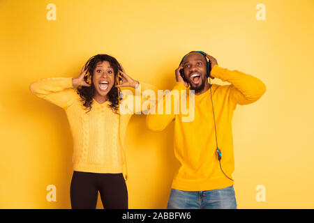 Young emotional african-american man and woman in bright casual clothes posing on yellow background. Beautiful couple. Concept of human emotions, facial expession, relations, ad. Listen to music, sing. Stock Photo