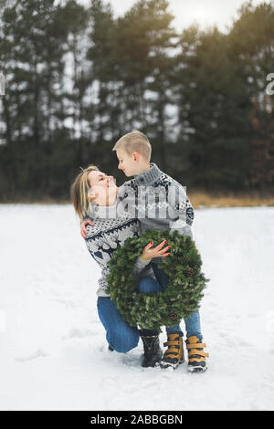 Merry Christmas and Happy Holidays. Mother and her little son walking together in winter forest and holding Christmas wreath. They are looking to each Stock Photo