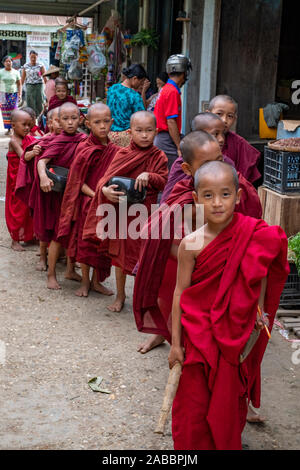 Many young Buddhist monks dressed in scarlet robes enter the market of Kanne, Myanmar (Burma) in search of alms and led by a young monk with a gong Stock Photo