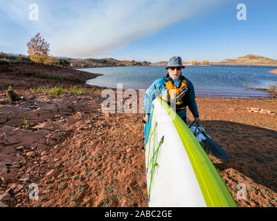 Senior male paddler is carrying stand up paddleboard on a lake shore in COlorado foothills, POV from action camera in a fall scenery Stock Photo