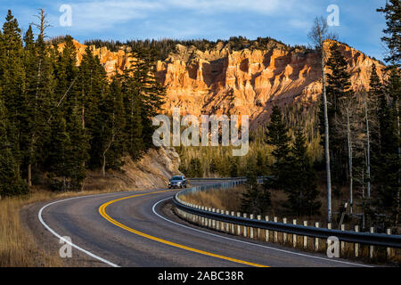 A winding road with silver minivan or SUV driving at sunset, below massive cliffs of red rocks and towers of sandstone in the Utah desert. Stock Photo