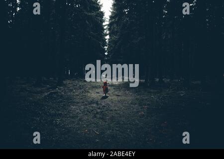 Little child with a red jacket stands in a dark forest