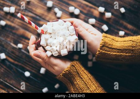 Female hands holding hot chocolate mug covered with marshmallow on dark wood background Stock Photo