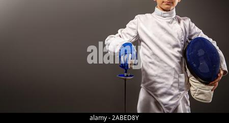 little boy fencer armed with a rapier in white sports equipment and with a protective helmet on a dark background Stock Photo