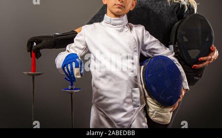 athlete and trainer mother and son in white traditional and black coaching protective fencing equipment with rapiers on a dark background Stock Photo