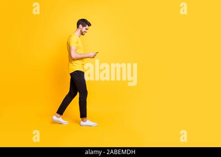Full length body size photo of cheerful attractive man with bristle smiling toothily focused on reading feednews on his phone wearing yellow t-shirt Stock Photo