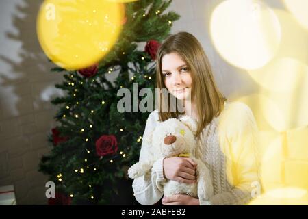 girl sits near a Christmas tree and holds in her hands a toy teddy bear Stock Photo