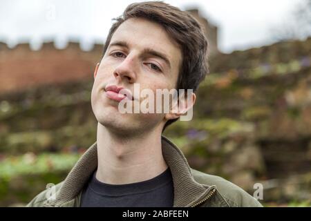 A confident handsome young man in his late teens or early twenties stands outdoors and looks to the camera with a questioning unconvinced expression. Stock Photo