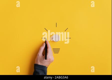Male hand drawing a light bulb with black marker with wooden cut circle as the bulb in the middle. Over yellow background with copy space. Stock Photo