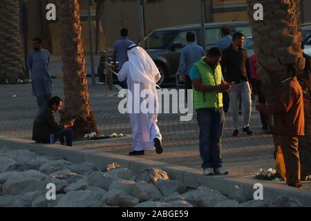 Kuwaiti man in traditional white robe (dishdasha) talking on the phone with Asian expatriates around him on the sidewalk Stock Photo