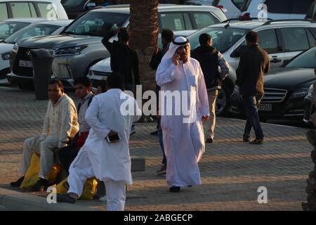 Kuwaiti man in traditional white robe (dishdasha) talking on the phone with Asian expatriates around him on the sidewalk Stock Photo