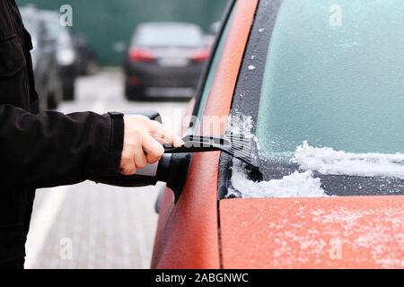 Man clears snow from icy windshield of car. Scraper in mans hand. Cleaning frozen window of orange automobile. Stock Photo