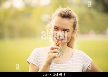 Woman eating a protein bar after outdoor workout - Closeup face of young blonde sporty woman resting while biting a nutritive bar Stock Photo