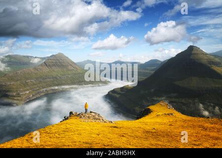 Lonely tourist in yellow jacket looking over majestic foggy fjords of Funningur, Eysturoy island, Faroe Islands. Landscape photography
