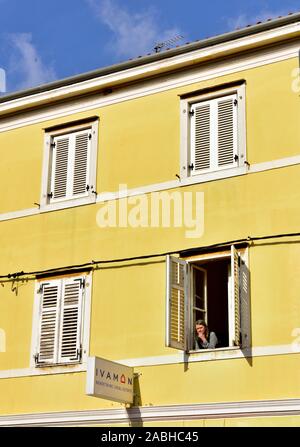 Looking up from outside at woman looking out of open window with shutters in yellow building Stock Photo
