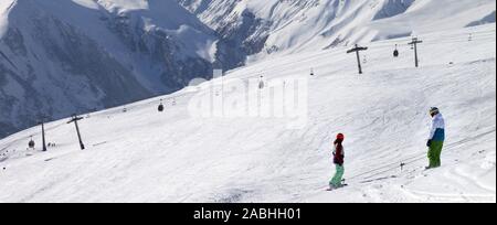 Two snowboarders descend on snowy ski slope and gondola lift at sun cloudy day. Georgia, region Gudauri. Caucasus Mountains in winter. Panoramic view. Stock Photo