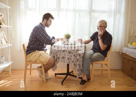 Young man and elder old man playing chess board game together at home. Stock Photo
