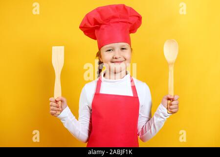 Beautiful chef child holds in his hands a wooden spoon and a spatula. Little girl in a cap and apron on a yellow background. Stock Photo