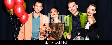 panoramic shot of cheerful multicultural women and handsome men holding champagne glasses near balloons on dark blue Stock Photo