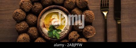 top view of falafel balls with hummus on plate near cutlery on wooden table, panoramic shot Stock Photo