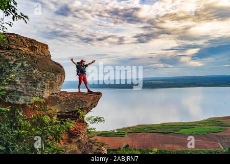 cheerful hiker man gesture raised arms on the edge of cliff, on a top of the rock mountain Stock Photo