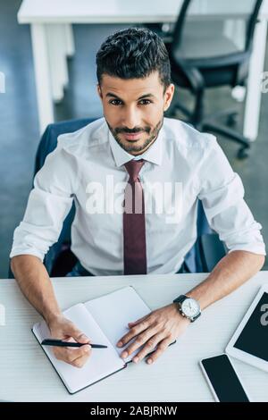 handsome businessman smiling at camera while writing in notebook at workplace Stock Photo