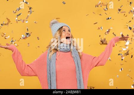 Shocked woman in hat and scarf with falling golden confetti isolated on yellow Stock Photo