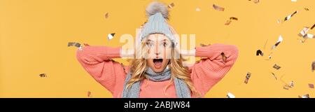 Shocked woman in hat with falling golden confetti isolated on yellow, panoramic shot Stock Photo