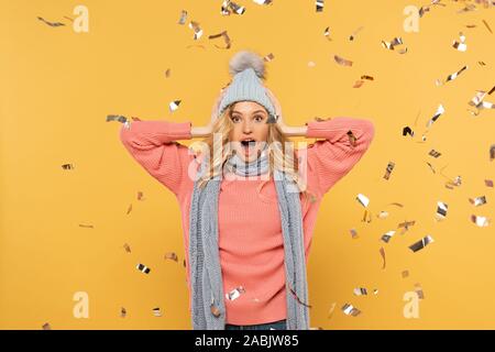 Surprised woman in hat and scarf with falling golden confetti isolated on yellow Stock Photo