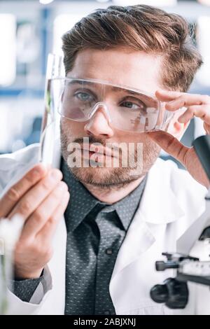 selective focus of handsome biochemist touching goggles and looking a test tube Stock Photo