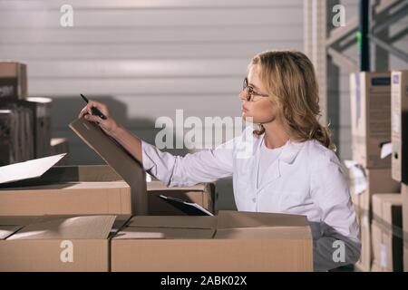 focused storekeeper in white coat inspecting cardboard boxes in warehouse Stock Photo