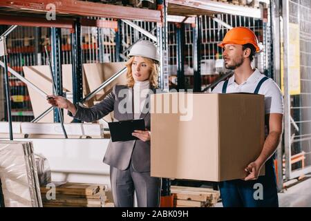 businesswoman in helmet pointing with hand while standing near loader holding cardboard box Stock Photo