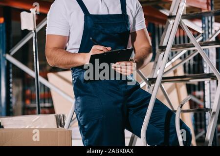 cropped view of warehouse worker in overalls writing on clipboard in warehouse Stock Photo