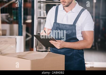 cropped view of warehouse worker writing on clipboard while standing near cardboard box Stock Photo