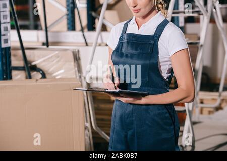 cropped view of workwoman in overalls writing on clipboard in warehouse Stock Photo