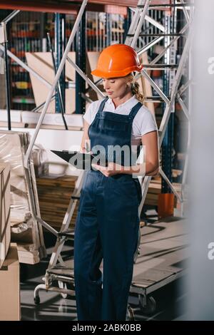 selective focus of workwoman in overalls and helmet standing near construction materials and writing on clipboard Stock Photo