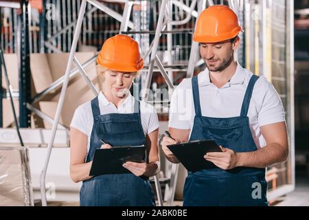 positive warehouse workers in overalls and helmets writing on clipboards Stock Photo