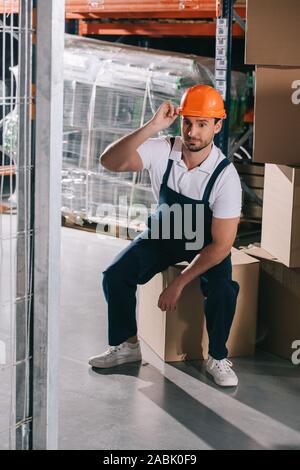 tired loader sitting on cardboard box in wearhouse and touching helmet Stock Photo