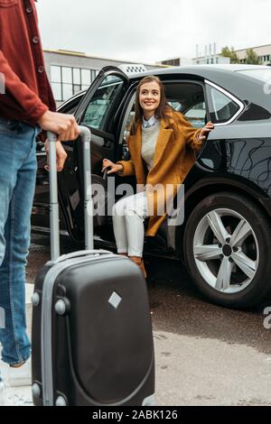 Driver holding wheeled suitcase and smiling woman sitting in taxi Stock Photo