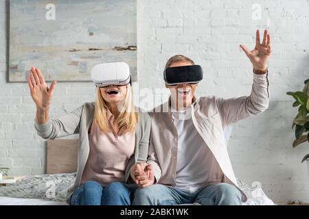 Exited couple in virtual reality headsets holding hands on bed Stock Photo