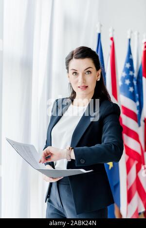 attractive diplomat holding folder and looking at camera near flags Stock Photo